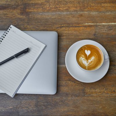A closed laptop, open notebook, and latte in a mug on a wooden surface.