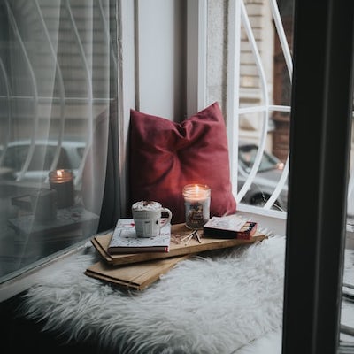 A cozy window bench with a blanket, pillow, and a candle, mug of hot chocolate, and book on a piece of wood.