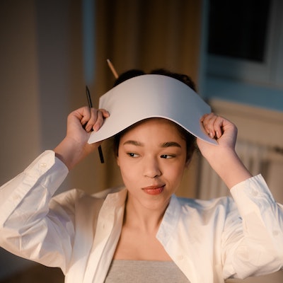A young woman holding a thin stack of papers up over her head with a pen in hand and a pensive smirk on her face.